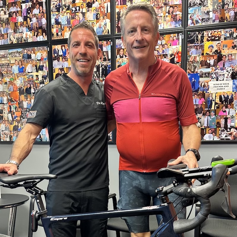 Dr Ryan Alter and a man stand beside a bicycle in front of a wall covered with photos